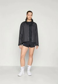 Giacca leggera nera con zip frontale, indossata sopra una maglietta nera e pantaloni corti. Sneakers bianche e calze con logo completano l'outfit.