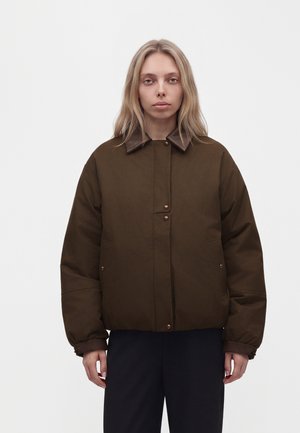 Une femme porte une veste matelassée marron foncé avec un col en cuir et des boutons-pression, debout devant un fond clair uni.