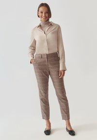 Beige saténová košile s knoflíky v kombinaci s hnědými kostkovanými kalhotami na míru. Modelka má na sobě černé baleríny. Hladká látka, strukturovaný střih a elegantní design.