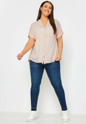 Blusa corta de manga corta en color rosa claro con botones en el frente, combinada con jeans ajustados de color azul oscuro y zapatillas blancas. Tela suave, ajuste relajado.