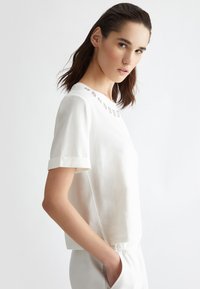 Top bianco a maniche corte con scollatura rotonda, dotato di risvolti e elementi decorativi sul colletto. Tessuto dalla consistenza liscia.