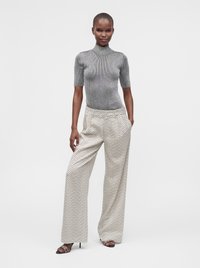 Femme portant un col roulé gris à manches courtes et un pantalon blanc large à motifs, debout avec une main dans la poche et portant des talons ouverts.