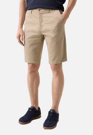 Man draagt beige knielange korte broek, donkerblauwe sneakers en een donkerblauw shirt, staat met handen in de zakken.