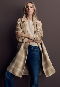 Cappotto lungo a quadri beige con colletto a rever, tessuto morbido e maniche larghe, indossato sopra un maglione leggero e jeans blu su uno sfondo strutturato.