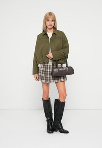 Veste verte courte, haut blanc, jupe mini à carreaux, et bottes noires jusqu'aux genoux. Sac à main en cuir marron avec une petite anse.