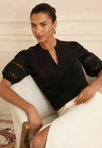 Blusa nera con scollo a V, maniche svasate con ritagli in pizzo, abbinata a una gonna color crema. Tessuto liscio, design minimalista.