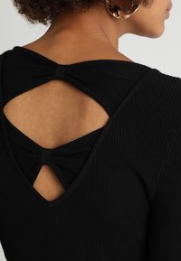 Maglione nero a coste con una profonda scollatura a V sul retro, decorato con due fiocchi in tessuto. La texture morbida e il design aderente mettono in risalto la scollatura sul retro.