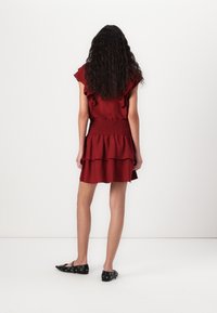 Femme aux longs cheveux bouclés portant une courte robe rouge à volants, debout face à un mur blanc et des chaussures plates noires avec des œillets.
