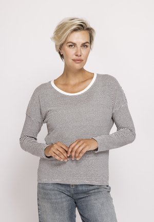 Blonde woman with short hair wearing a black-and-white striped long-sleeve shirt and blue jeans, standing with hands clasped.