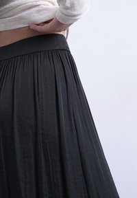 Άτομο που σηκώνει ελαφρώς λευκό πουκάμισο για να αποκαλύψει τη μέση, φοράει μαύρη πλισέ φούστα ψηλόμεση, σε απλό λευκό φόντο.