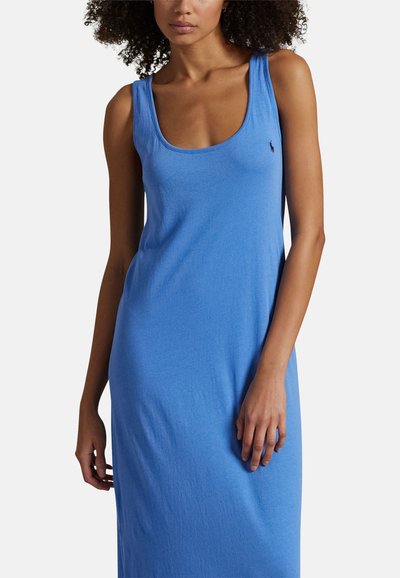 Kobieta o kręconych włosach, ubrana w niebieską sukienkę na ramiączkach z dekoltem w serek i małym haftowanym logo na piersi, na białym tle.