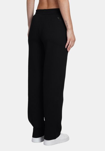 Zwarte trainingsbroek met een relaxte pasvorm, elastische tailleband en zijzakken. Opvallend logo detail aan de achterkant, gecombineerd met witte sneakers.