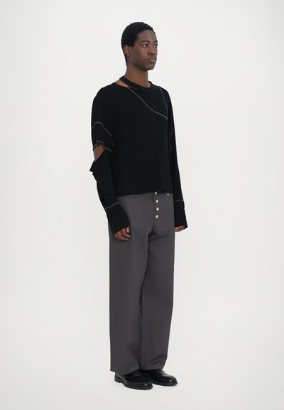 Schwarzes Langarmshirt mit Cut-out-Details und grauen Nähten, kombiniert mit grauen weiten Hosen mit Knopf-Akzenten. Schwarze Schuhe.