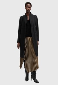 Cappotto lungo nero su una maglietta nera con texture, abbinato a una gonna plissettata dorata e stivali neri con tacco al ginocchio. Sfondo neutro.