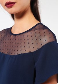 Mekko navy-värissä, jossa on läpikuultava pisteverkko yoke, pyöreä kaula-aukko ja lyhyet liehuvat hihat. Silkkinen kangas, jossa on tekstuuritietoa olkapäällä.