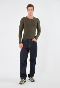 Oliven langermet tettsittende skjorte kombinert med løse mørkeblå jeans. Modellen har på seg grønne og svarte mønstrede joggesko. Enkel hvit bakgrunn.