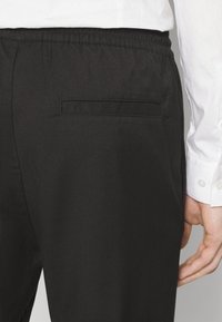Pantalon noir avec une taille élastique, doté d'une poche arrière et d'une texture lisse, porté avec une chemise de couleur claire.