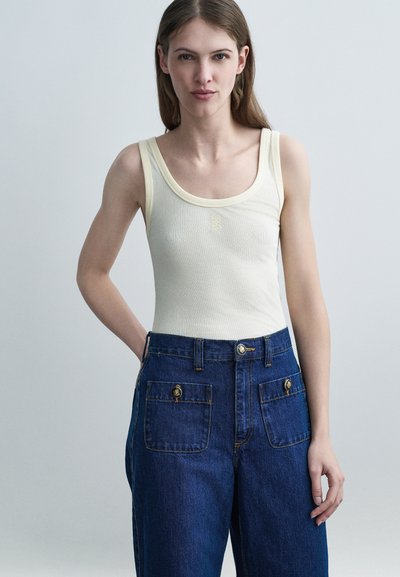 Jeune femme aux longs cheveux bruns portant un débardeur crème côtelé et un jean bleu foncé taille haute à jambes larges avec poches avant.