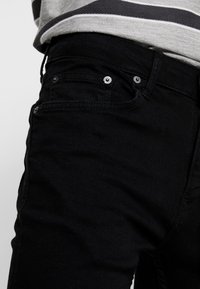 Jeans de mezclilla negra con diseño de cinco bolsillos, herrajes metálicos visibles y una textura suave. La tela parece ligeramente elástica.