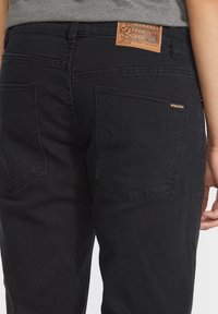 Black jeans with a fitted design, featuring a leather patch on the waistband, two back pockets, and a subtle logo accent.