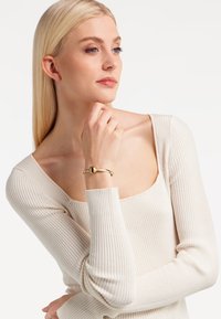 Brățară de culoare aurie cu un accent sferic neted, expusă pe un încheietură. Persoana poartă un top alb cu dungi, cu mânecă lungă.