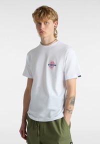 T-shirt bianca in cotone con un logo Vans blu e rosso sul petto, maniche corte e collo rotondo classico.