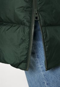 Mørkegrøn polstret jakke delvist åbnet, der afslører blå denimjeans underneath.