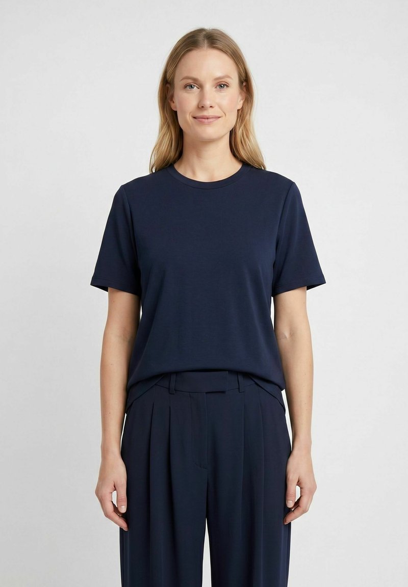 Femme aux cheveux blonds mi-longs, portant une chemise bleu marine à manches courtes et un pantalon plissé assorti, debout devant un fond uni.