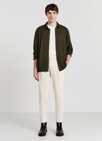 Camicia button-up verde oliva sopra una maglietta bianca, abbinata a pantaloni color crema e stivaletti neri alla caviglia. Tessuti lisci e design aderente.