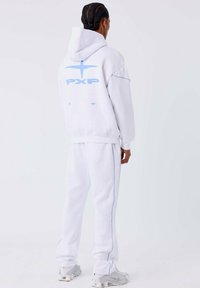 Persoon met gevlochten haar, gekleed in een witte hoodie en bijpassende sweatpants met lichtblauwe details, staand tegen een effen witte achtergrond.