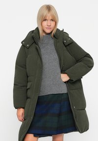 Olivenfarvet kordfløj pufferjakke med aftagelig hætte, trykknapper og sidelommer, båret over en grå sweater og en ternet nederdel.