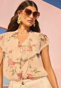 Blusa floreale in tessuto leggero e trasparente con bordi arricciati, caratterizzata da motivi rosa, blu e verdi; abbinata a occhiali da sole oversize marroni e orecchini d'oro.