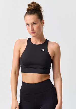 Frau mit hellem Teint und einem Dutt, trägt einen schwarzen Sport-BH mit hohem Kragen und passende hoch taillierte Leggings, steht vor einem einfarbigen grauen Hintergrund.