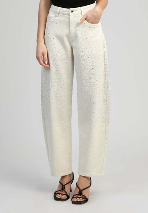 Femme portant un jean blanc taille haute orné de petits strass et des sandales à talons noires à brides, debout avec une main dans la poche.