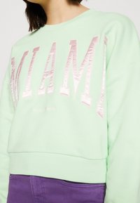 Felpa verde menta con la scritta "MIAMI" in rosa pallido lucido. Presenta un design accorciato e polsini a costine, abbinata a pantaloni viola.
