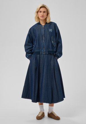 Femme aux cheveux blonds courts portant une veste en jean foncée et une longue jupe en jean, debout les mains dans les poches devant un fond uni.