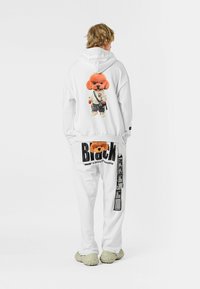 Persona vestida con sudadera blanca y pantalones con un gráfico de perro naranja en la parte trasera y el texto negro en negrita "Black" en los pantalones, contra un fondo blanco.