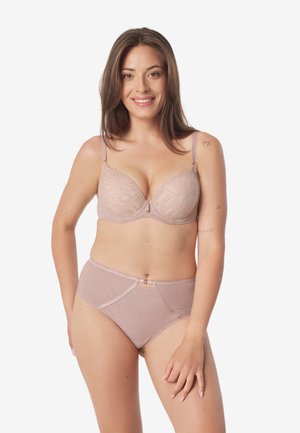 Kobieta o długich brązowych włosach, ubrana w beżowy biustonosz i wysokie majtki z kwiatowym wzorem, uśmiechająca się z ręką spoczywającą na udzie.