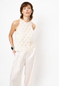 Crème mouwloos topje met een gestructureerd, geschulpte ontwerp; gecombineerd met losse lichte broeken. Glad materiaal met subtiele glans, minimale details.