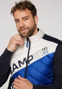 Gilet imbottito bianco e blu con manica blu navy. Caratterizzato da un alto colletto, chiusura con zip e un audace testo "CAMP 01 BLUE". Motivo a rombi imbottito.