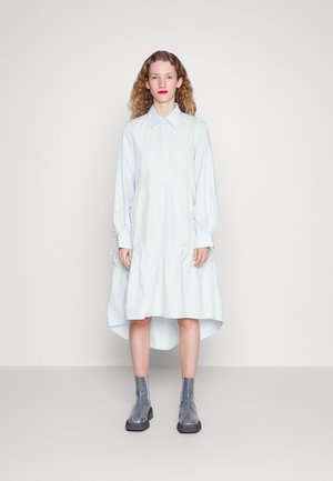 Lyseblå, langermet kjole med krage, lagdelt hem og lett hevet bak. Kombinert med grå ankelstøvletter og minimalistiske designdetaljer.