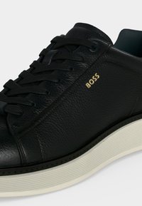 Baskets en cuir noir avec lacets noirs, semelle blanche texturée et petit logo doré "BOSS" sur le côté.