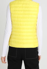 Gelbe Pufferweste mit hohem Kragen, vertikaler Naht und leichtem, gestepptem Design. Getragen über einem langärmligen, weißen Shirt.