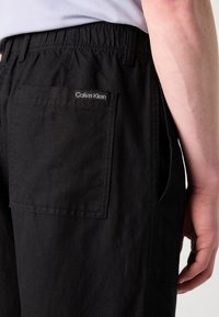 Black elastic-waist pants made of lightweight fabric, featuring a back pocket with a white logo label. Smooth texture, relaxed fit.