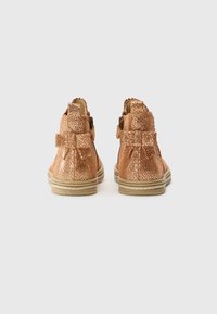 Botas de tobillo de cuero bronce con un acabado texturizado, que presentan un lazo en la parte trasera y una suela de color claro.