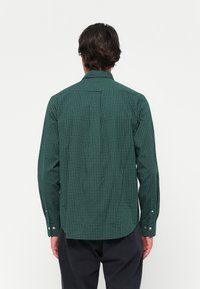 Cămașă verde cu un model, cu un design punctat deschis, mâneci lungi și mansete cu nasturi. Prezintă un guler obișnuit și o tivă curbată.