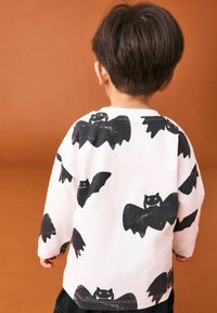 Camicia bianca a maniche lunghe con illustrazioni di pipistrelli neri, con scollatura rotonda e vestibilità comoda, su uno sfondo marrone.