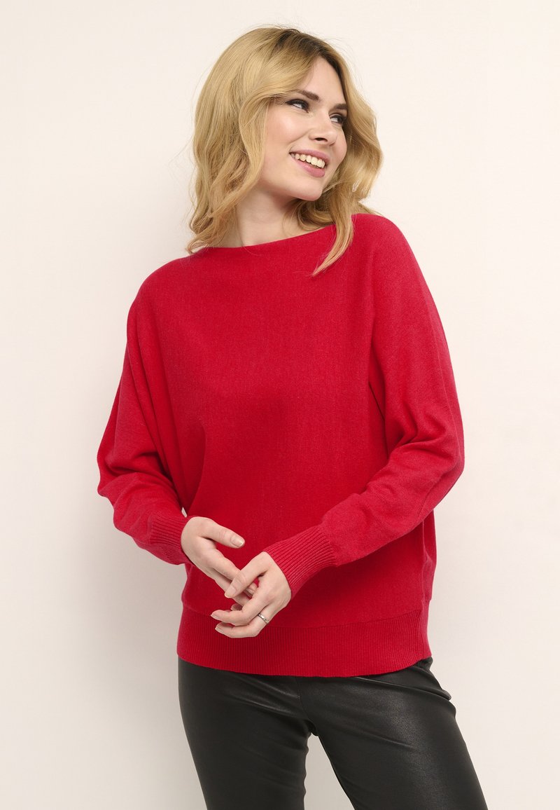 Maglione rosso oversize realizzato in tessuto morbido con polsini e orlo a costine. Indossato sopra pantaloni di pelle nera, con scollatura a barca.