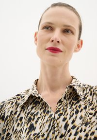 Tehénmintás blúz krém, fekete és arany színekben, gallérral és gombzáródással, sima, fényes anyagból készült.