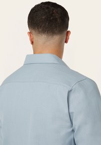 Lichtblauw, textuur shirt met een achterpaneel en kraag. De stof lijkt soepel en gestructureerd, wat een getailleerde pasvorm benadrukt.